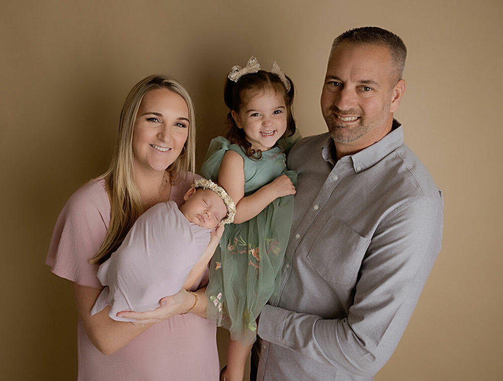 A family portrait of a man and woman smiling and posing with their two daughters during their pastel newborn session in Southampton, New Jersey.