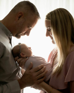 A close-up of a man and woman holding their baby girl in their arms as they both look at her. They are standing across from each other against a light and bright set during their pastel newborn session in Eastampton, New Jersey.