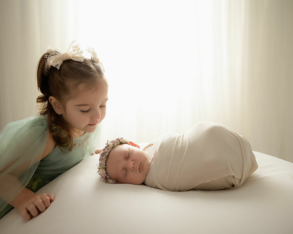A sibling portrait of a toddler girl looking at her sleeping baby sister sleeping on bean bag photography prop, wearing cute dress with bow and headbands against a light and bright backdrop in Westampton, New Jersey.