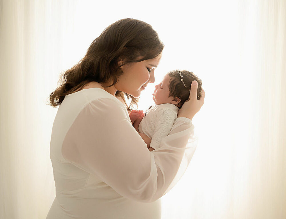 A cute mommy and me portrait of a woman holding her baby in her arms almost touching noses as she sleeps against a light and bright backdrop during her light pink newborn session in Southampton, New Jersey.