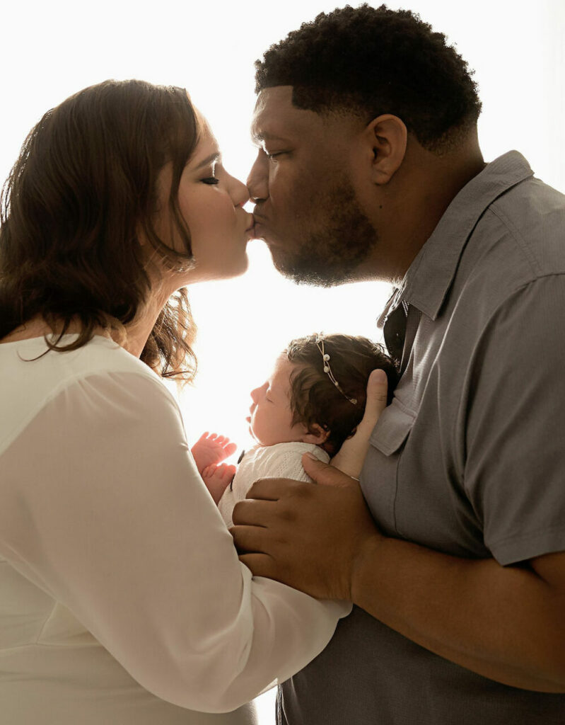 A family portrait of a man and woman kissing as they both had their baby girl in their arms I can see the light and bright backdrop for their daughters light pink newborn session in Eastampton, New Jersey.