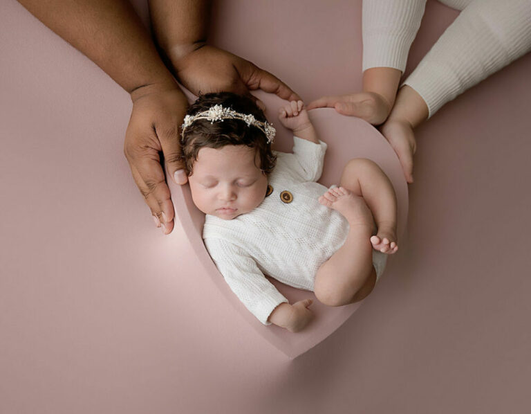 A beautiful infant portrait of a baby girl resting on heart shaped photography prop with her parents hands in the photo for her professional baby pics in Medford, New Jersey.