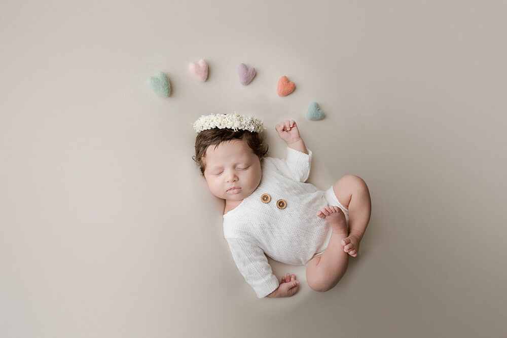 A cute portrait of a newborn girl sleeping on her back wearing cute outfit and flowery headband with felt hearts above her head sleeping during her light pink newborn session in Mount Holly, New Jersey.