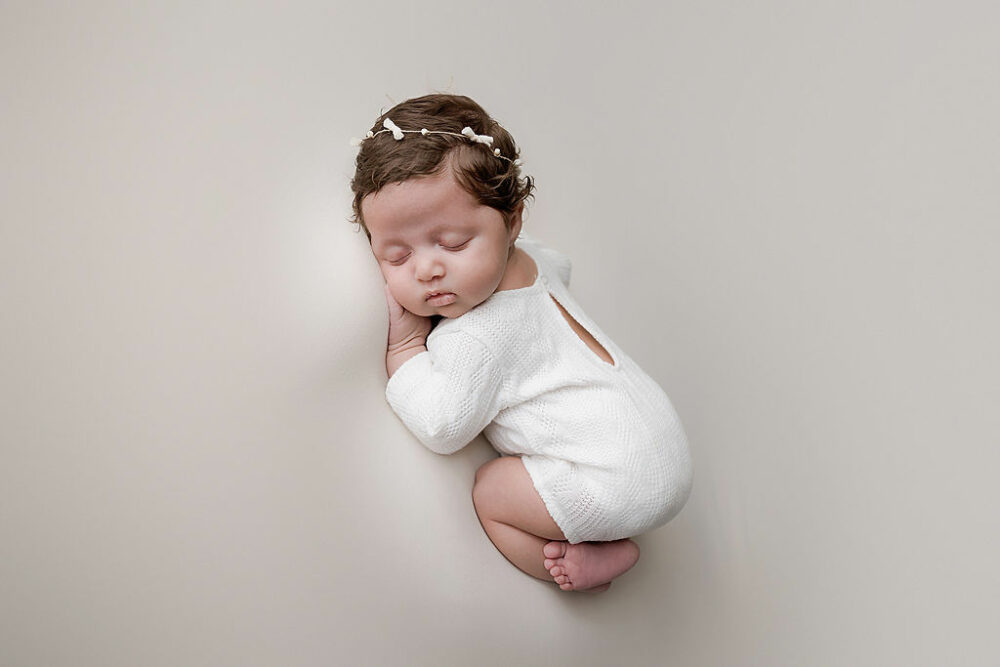 A close-up of a sleeping baby girl resting on her tummy wearing cute outfit and dainty headband sleeping during her light pink newborn session in Cherry Hill, New Jersey.