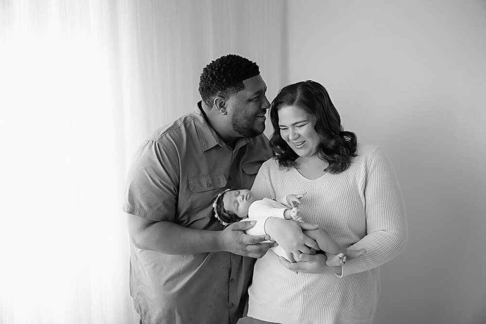 A family portrait of a man and a woman smiling and laughing during their daughters light pink newborn session in Pemberton, New Jersey.
