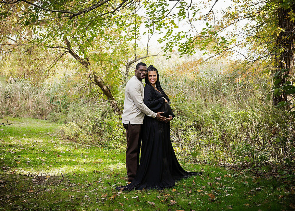 A man and woman doing classic prom pose as a both hold her belly for their cute maternity pictures taken outdoors for professional photography in Palmyra nature Cove, New Jersey.
