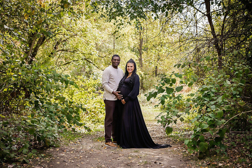 A couple portrait of a man and a woman standing together on a park trail posing in holding her belly for their couple pregnancy photos at Palmyra Nature Cove, New Jersey.