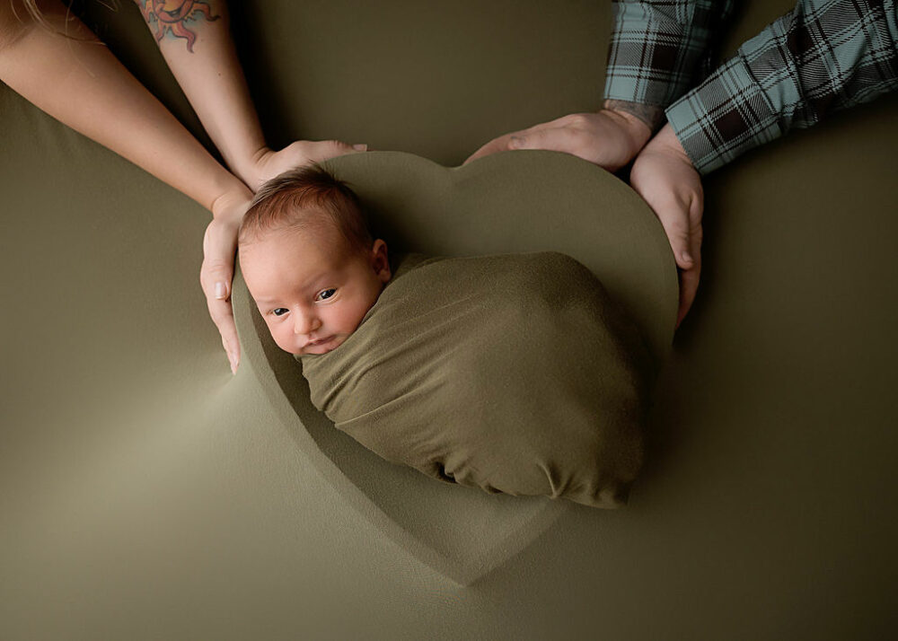 Cute newborn portrait of a baby awake and resting on its back in a heart shape photography prop with earthy colors during her newborn session in Delran, New Jersey.