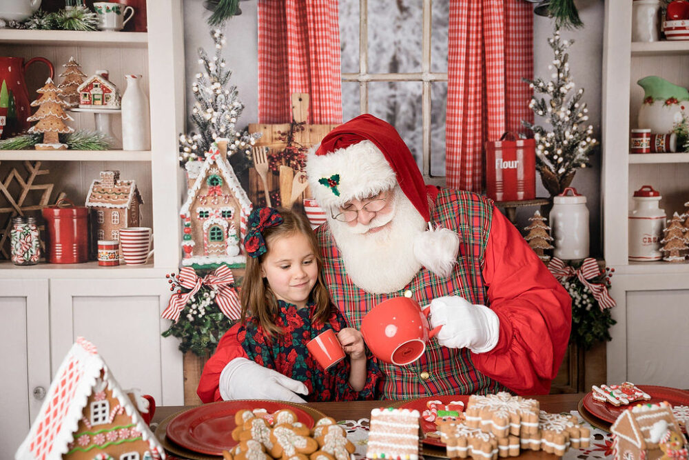 Photo of a toddler girl drinking hot cocoa with Santa Claus in his gingerbread kitchen set during Stinsman photography Christmas experience session in Pemberton, New Jersey.