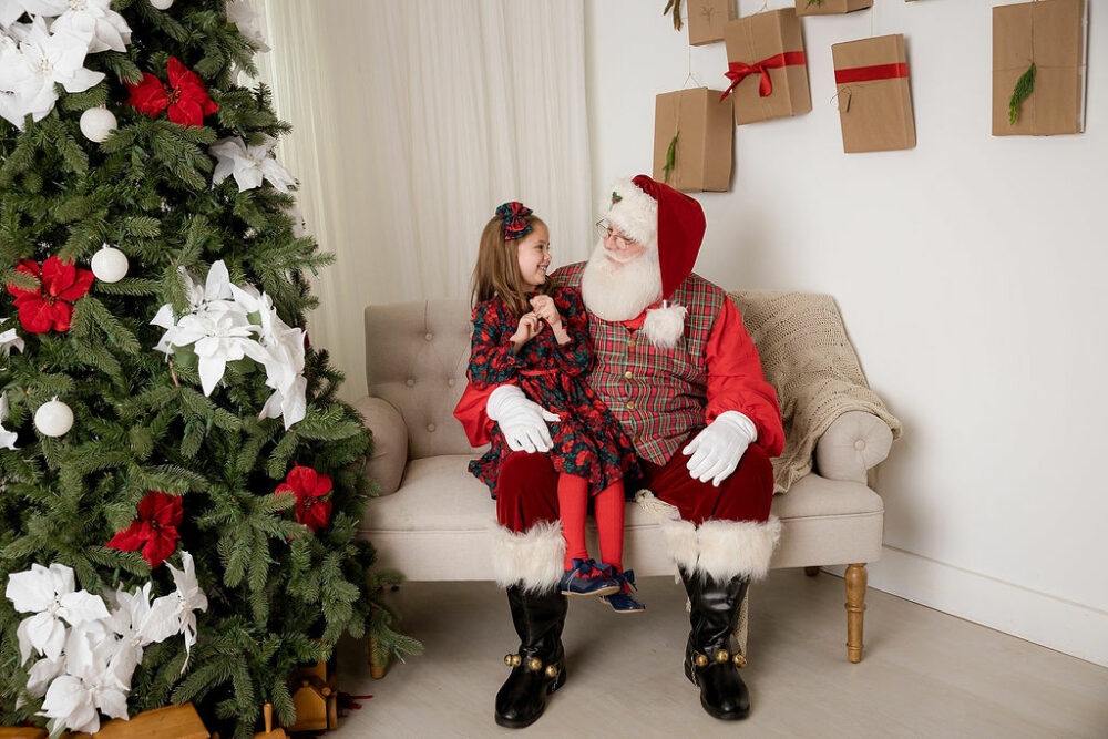 A photo of a toddler girl talking with Santa as they sit on sofa with large Christmas tree during Santa mini sessions in Cherry Hill, New Jersey.