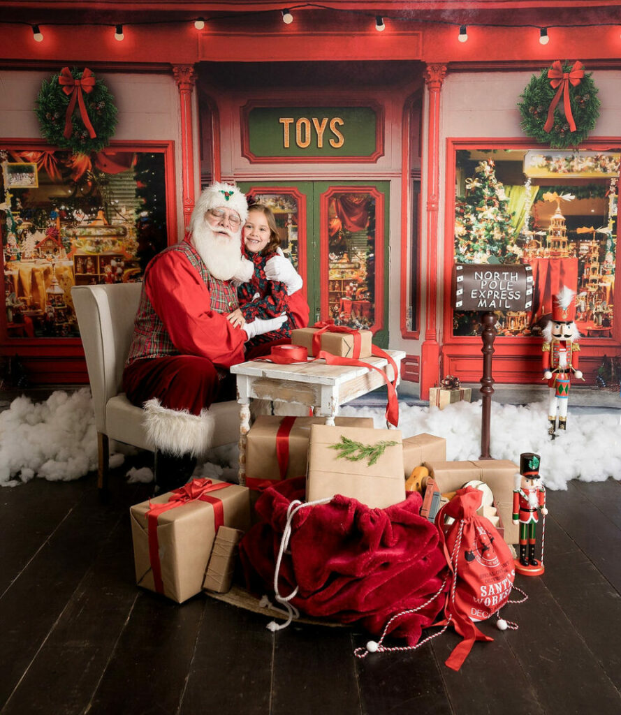 An adorable photo of a toddler girl hugging Santa Claus in his toy shop set for professional photography Santa minis in Hamilton, New Jersey.