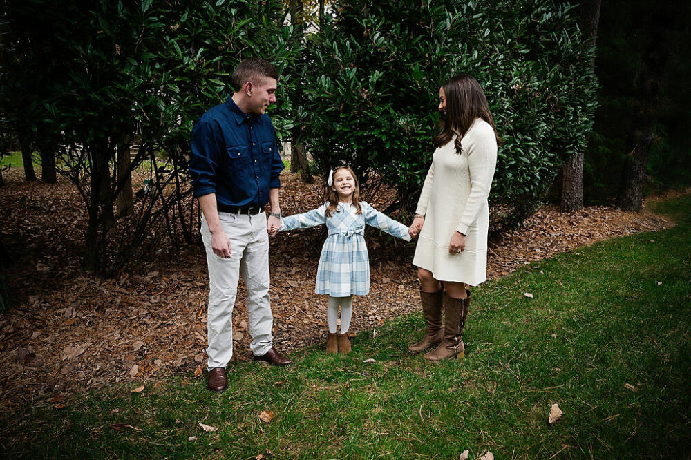 Family portrait of a man and woman holding their daughters hand as she smiles between them for their outdoor fall family session in Medford, New Jersey.