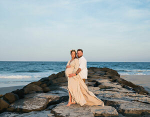 A beautiful couple maternity portrait of a man and woman standing on rocks at the beach posing for their professional pregnancy portraits in toms River, New Jersey.